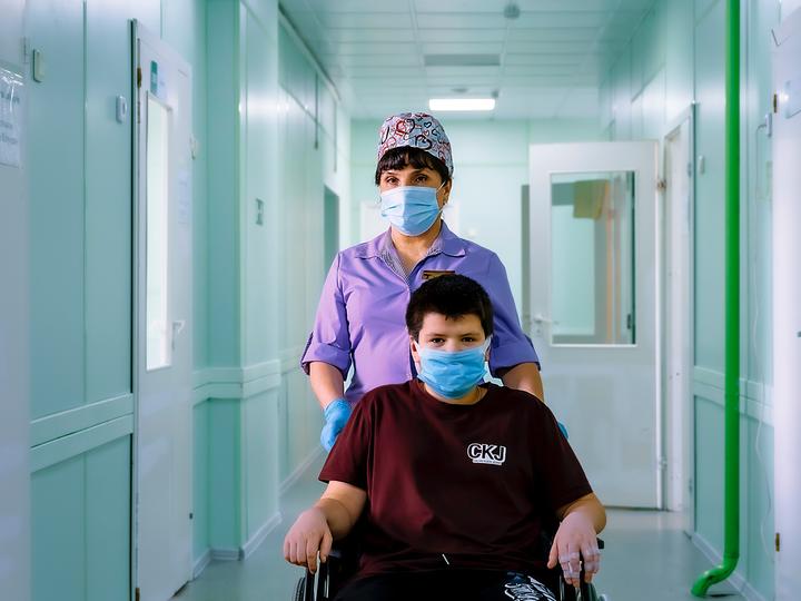 Nurse in purple scrubs and mask walking patient in mask and wheelchair down a facility hallway.